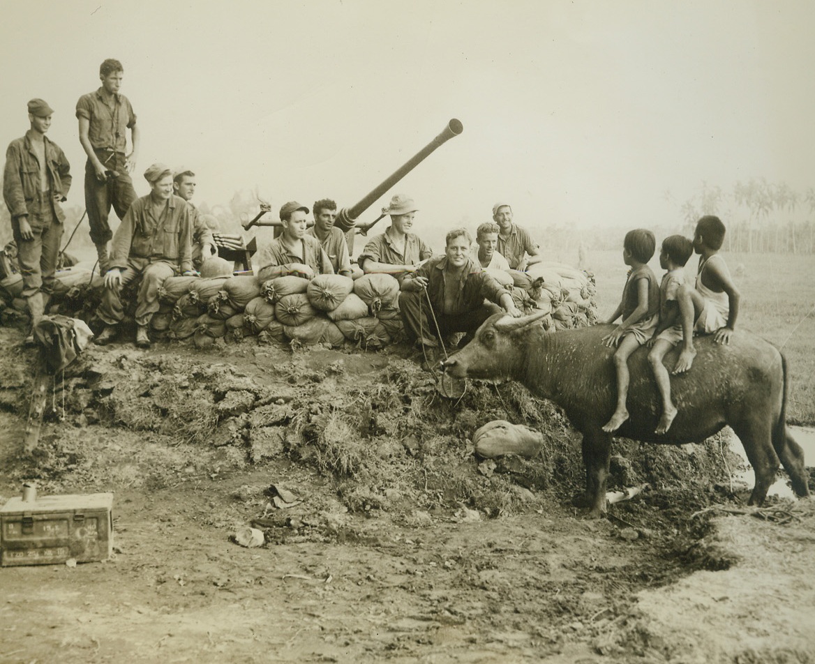 Yanks Find Out “What’s Cooking’”, 11/23/1944. Leyte Island, P.I. – During a lull in the fighting, members of a Yank ack-ack gun crew “shoot the bull” with three small citizens of Leyte island, perched atop their family’s patient caribou.  Photo by ACME photographer, Tom Shafer, for the War Picture Pool.Credit-WP-(ACME);