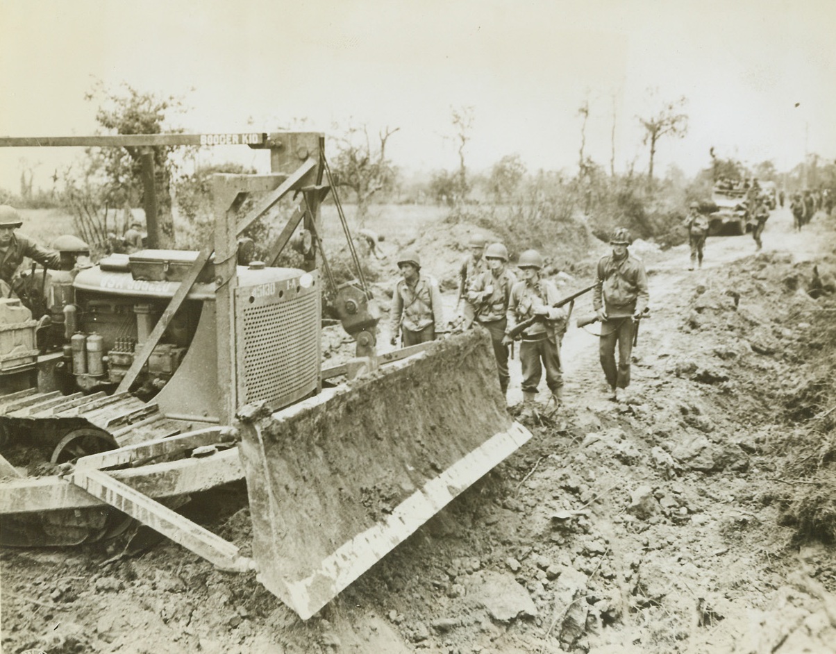 It Puts In and Takes Away, 7/29/1944. France – While trucks wait in the background, the mighty bulldozer fills in bomb craters on a road in France, to clear the way for advancing American forces. The versatile machine has also proved a great asset in clearing debris-filled streets in captured towns. Credit: Signal Corps photo from ACME;