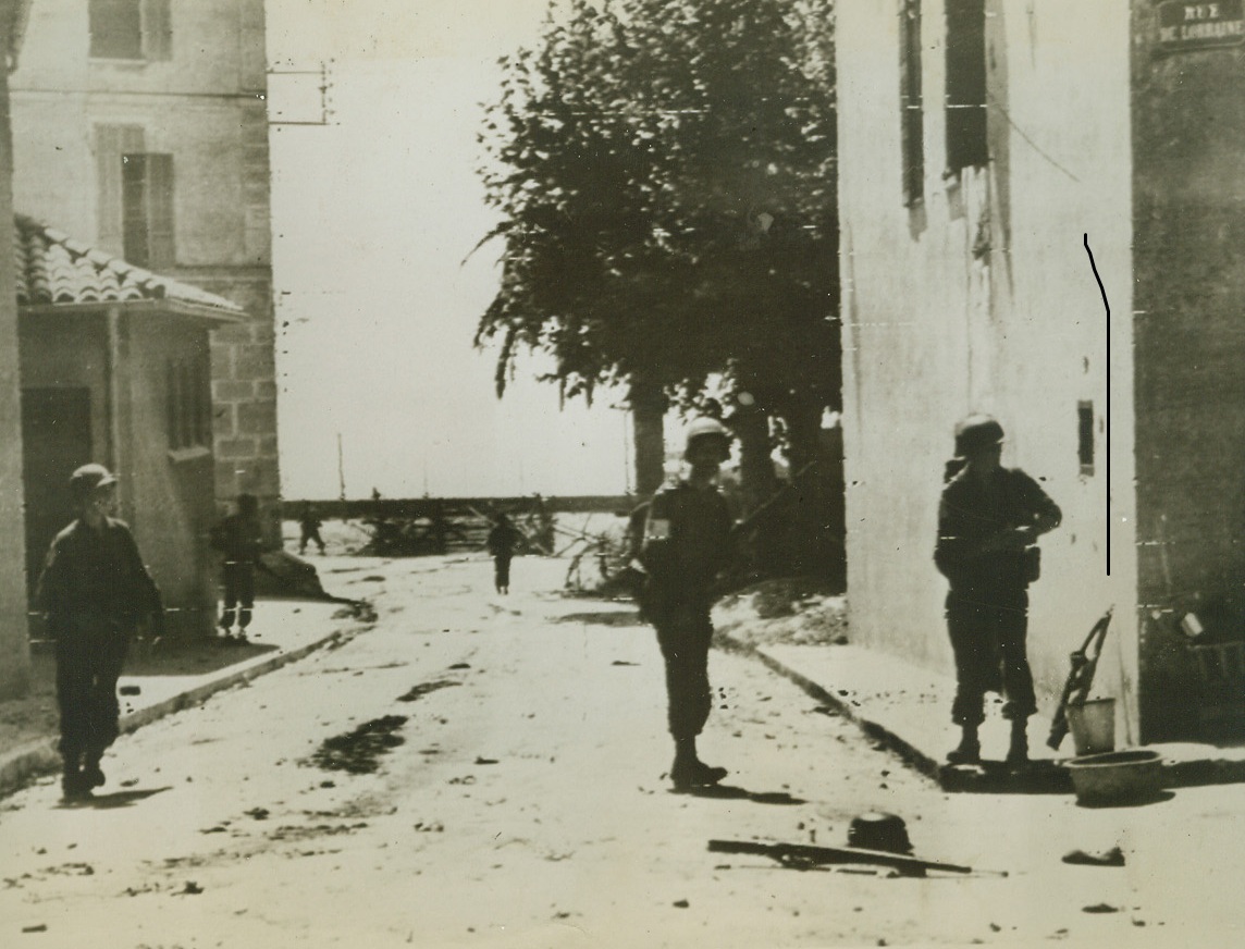 Expanding New Beachhead, 8/16/1944. Moving cautiously and watching every house for enemy snipers, American Infantrymen advance through a street in a town east of Toulon. In the background, can be seen the beach, one of the many points Allied invasion forces hit along the coast of Southern France. Note German rifle and helmet (right, foreground). Credit: ACME PHOTO BY SHERMAN MONTROSE FOR WAR PICTURE POOL VIA ARMY RADIOTELEPHOTO.;